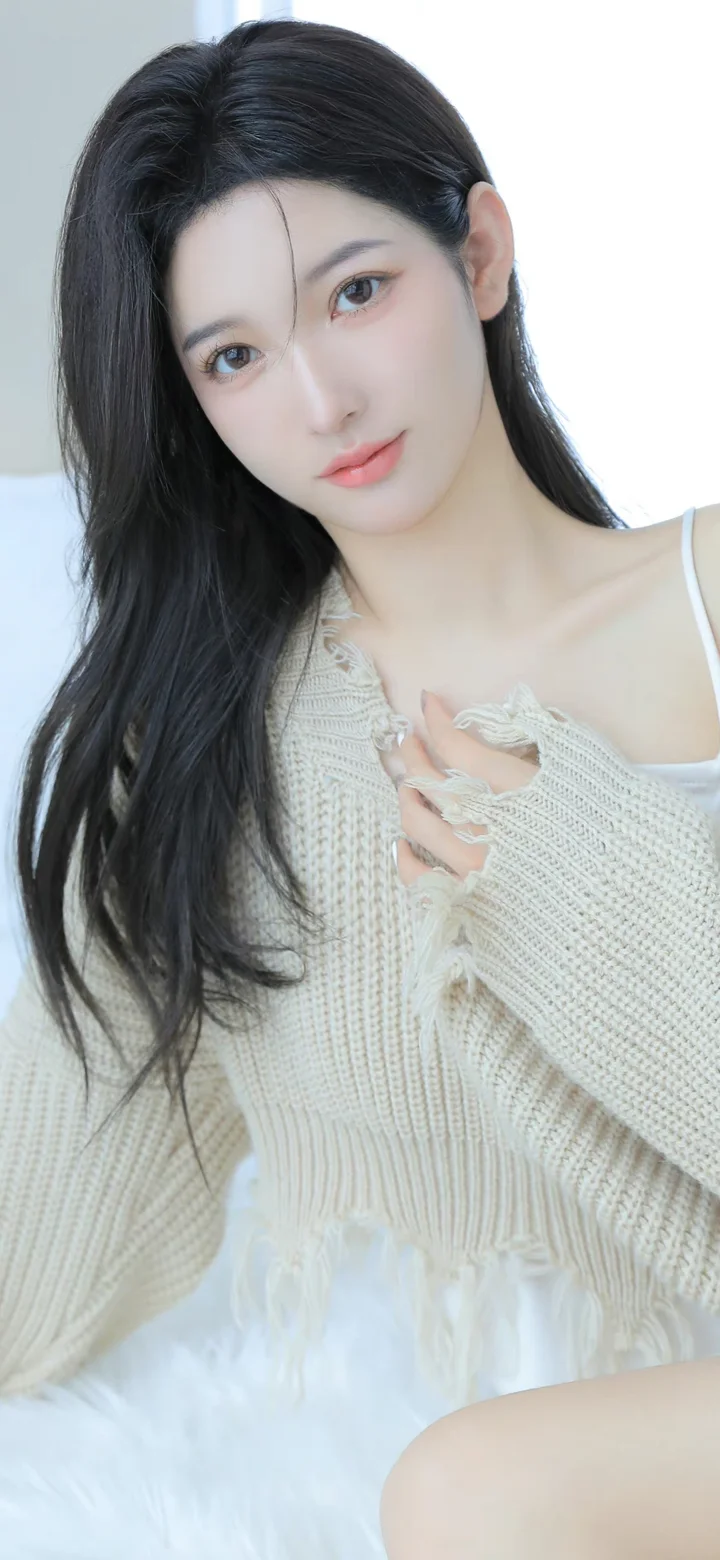 一名长发女子穿着白色针织衫坐在室内白色背景前。