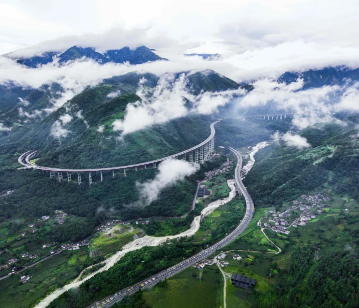 云雾笼罩的绿色山谷中，蜿蜒的高架公路、河流和村落清晰可见。