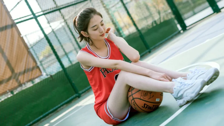 빨간 운동복을 입은 여성이 농구공 옆 농구 코트에 앉아 있습니다.