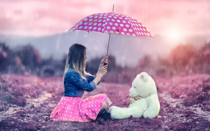 一名女子坐在地面上撑着粉色波点雨伞，身旁放着一只白色泰迪熊，周围可见雨景。