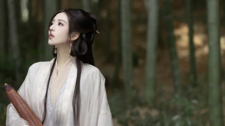 一名身穿浅色传统服饰的女子站在竹林中，抬头望向左上方。