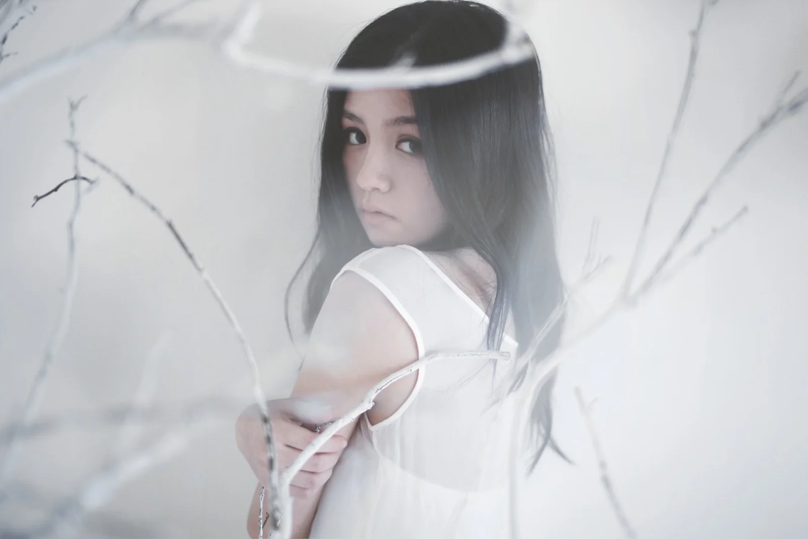 흰 옷을 입은 여성이 뒤돌아 카메라를 바라보고 옅은 나뭇가지가 전경을 가로지른다.