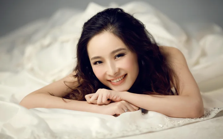 一名成年女性趴在白色床品上，微笑着将手臂交叠在身前。