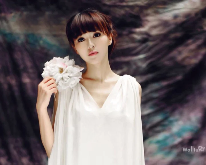 一名身穿白色服装的成年女子手持白花站在模糊的抽象背景前。