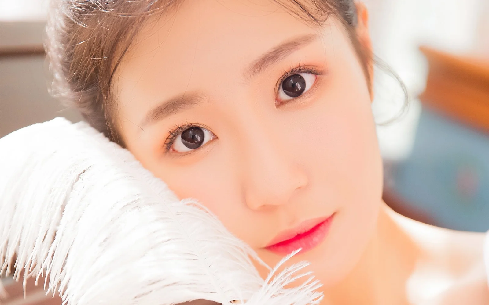 女性の顔のクローズアップが画面を占め、明るい瞳とピンク色の唇の横に白い羽毛のような質感が見える。