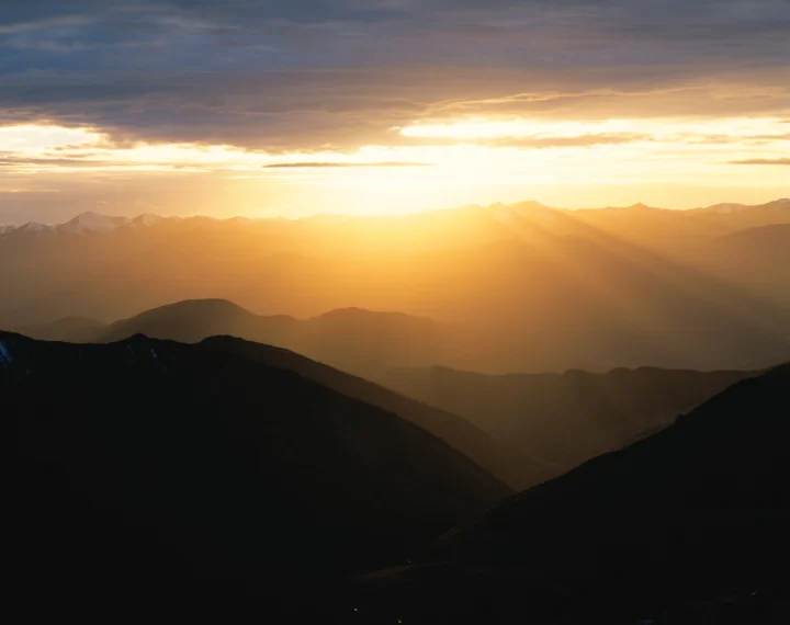夕阳穿过云层洒在层叠山脉之间，形成金色的山间暮色。