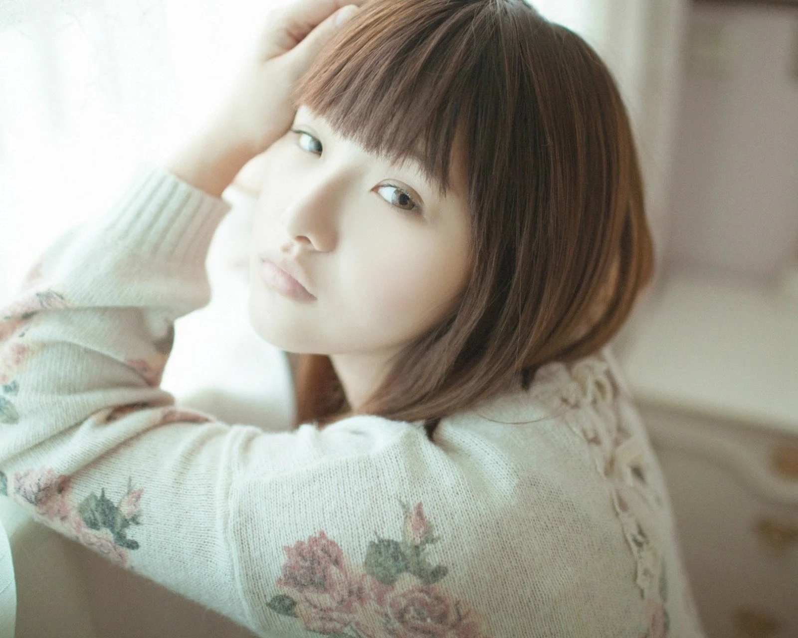 花柄の淡いニットを着たショートヘアの女性が、窓辺でカメラに目を向けている。