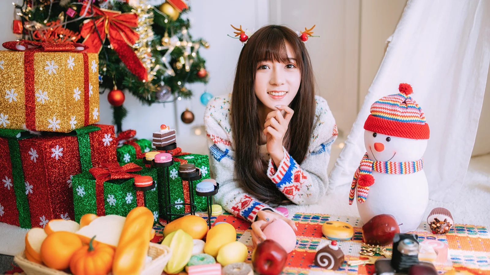 한 여성이 크리스마스트리와 선물 상자, 눈사람 옆에 앉아 있고 테이블에는 과일과 디저트가 놓여 있습니다.