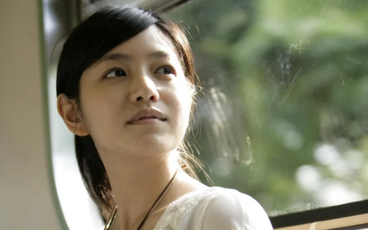 女性が窓辺に立ち、顔に陽光が差し込み、背景には柔らかな緑の景色が広がっています。