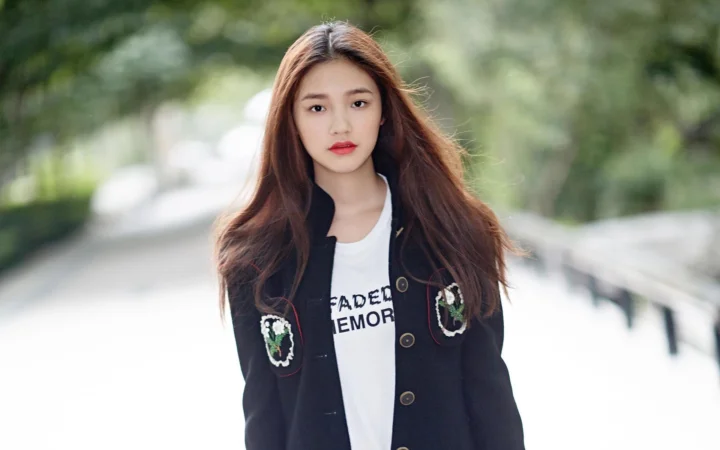 一名长发女性站在户外林荫道路旁，身穿黑色外套和白色上衣。