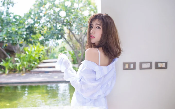 一名穿白色露肩上衣的女子站在花园泳池旁回望镜头。