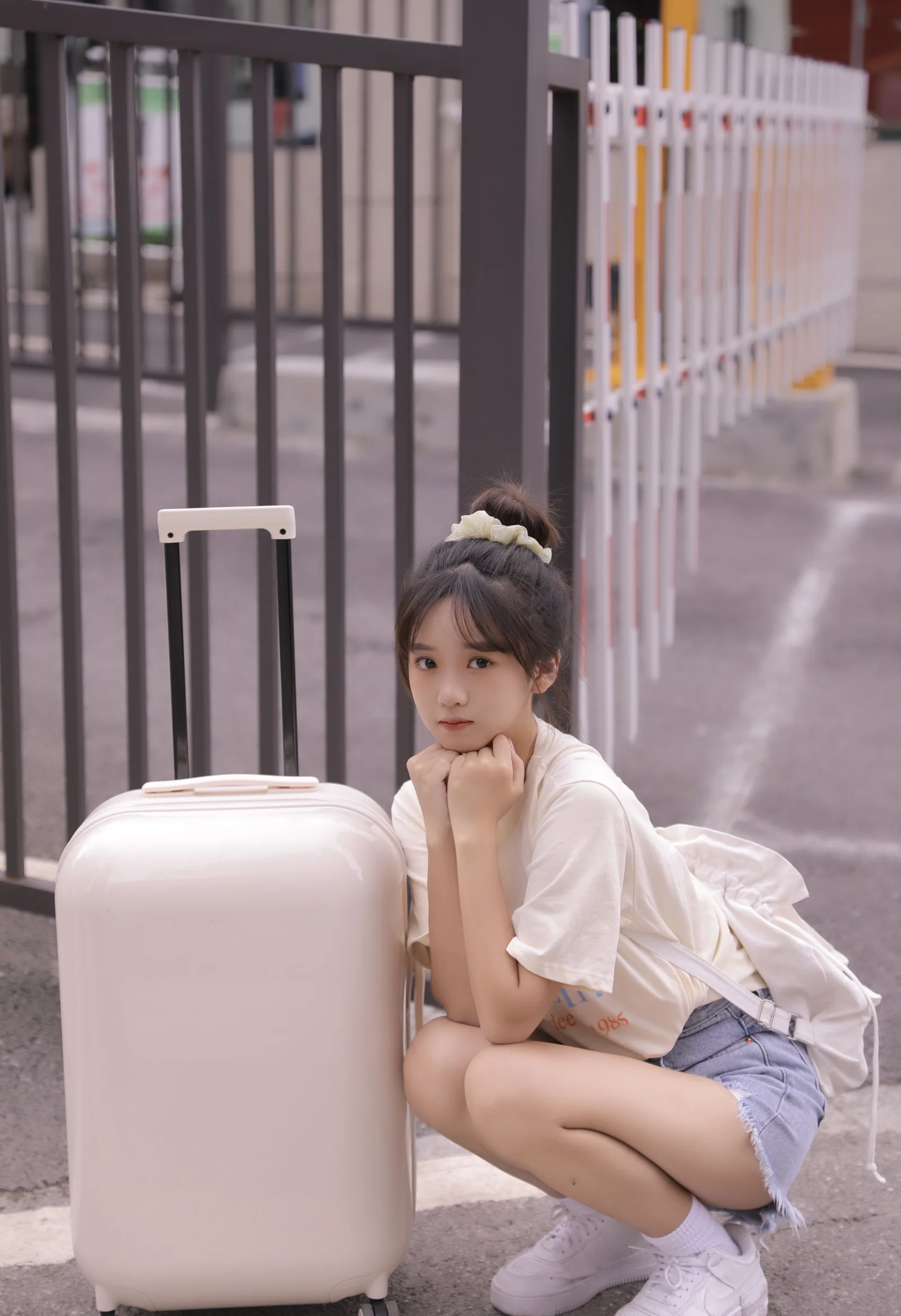 一名年轻女子蹲在户外栏杆旁的浅色行李箱边。
