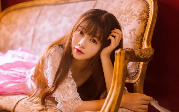 긴 머리의 여성이 화려한 나무 팔걸이 의자 곁에 기대어 있는 따뜻하고 부드러운 실내 인물 사진입니다.