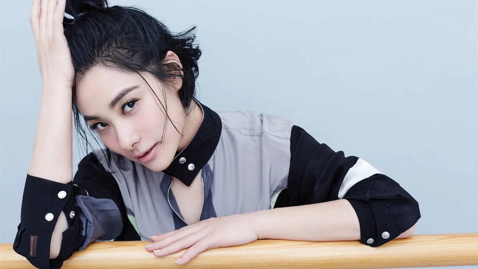 검정과 회색 상의를 입은 성인 여성이 나무 테이블에 기대어 카메라를 향해 미소 짓고 있다.
