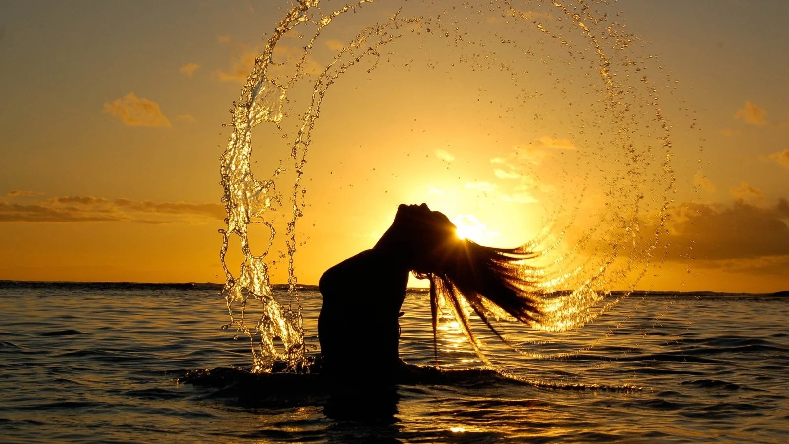 夕日に照らされ、長い髪の人物が水しぶきに囲まれて海の中に立っています。