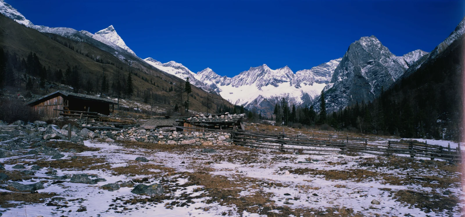 积雪覆盖的高山山谷在深蓝天空下延展开来，前景可见木屋和围栏。