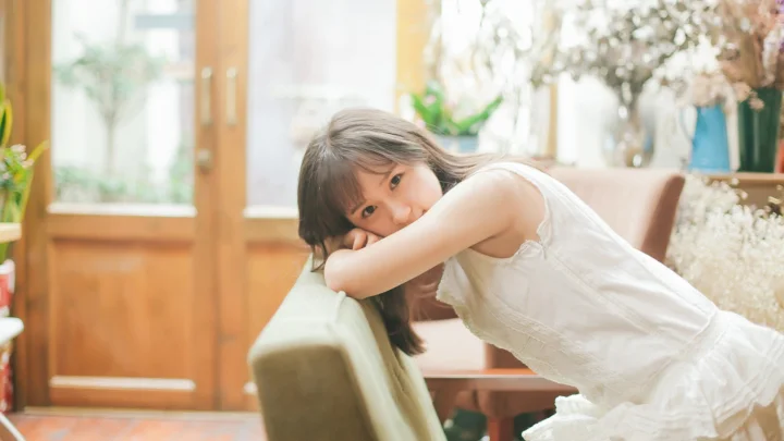 흰색 원피스를 입은 여성이 밝고 따뜻한 실내에서 의자 등받이에 기대고 있습니다.