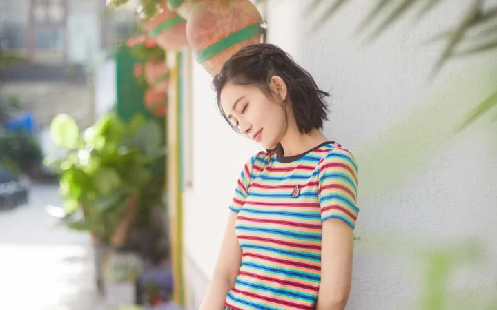 カラフルなボーダーシャツを着た女性が、屋外の白い壁にもたれて目を閉じています。