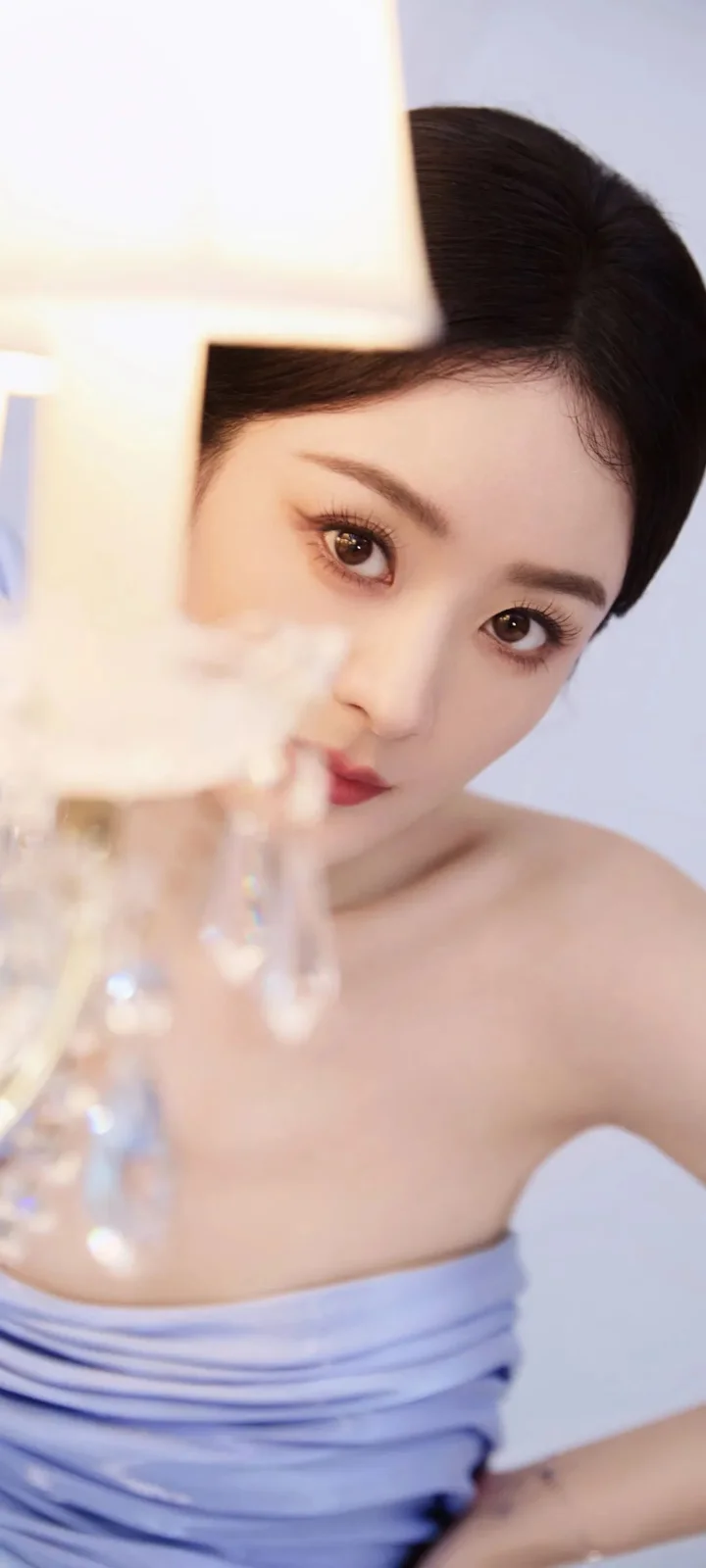一名成年女性身穿浅紫色无肩带上衣，在柔和光线和前景灯具的遮挡下看向镜头。