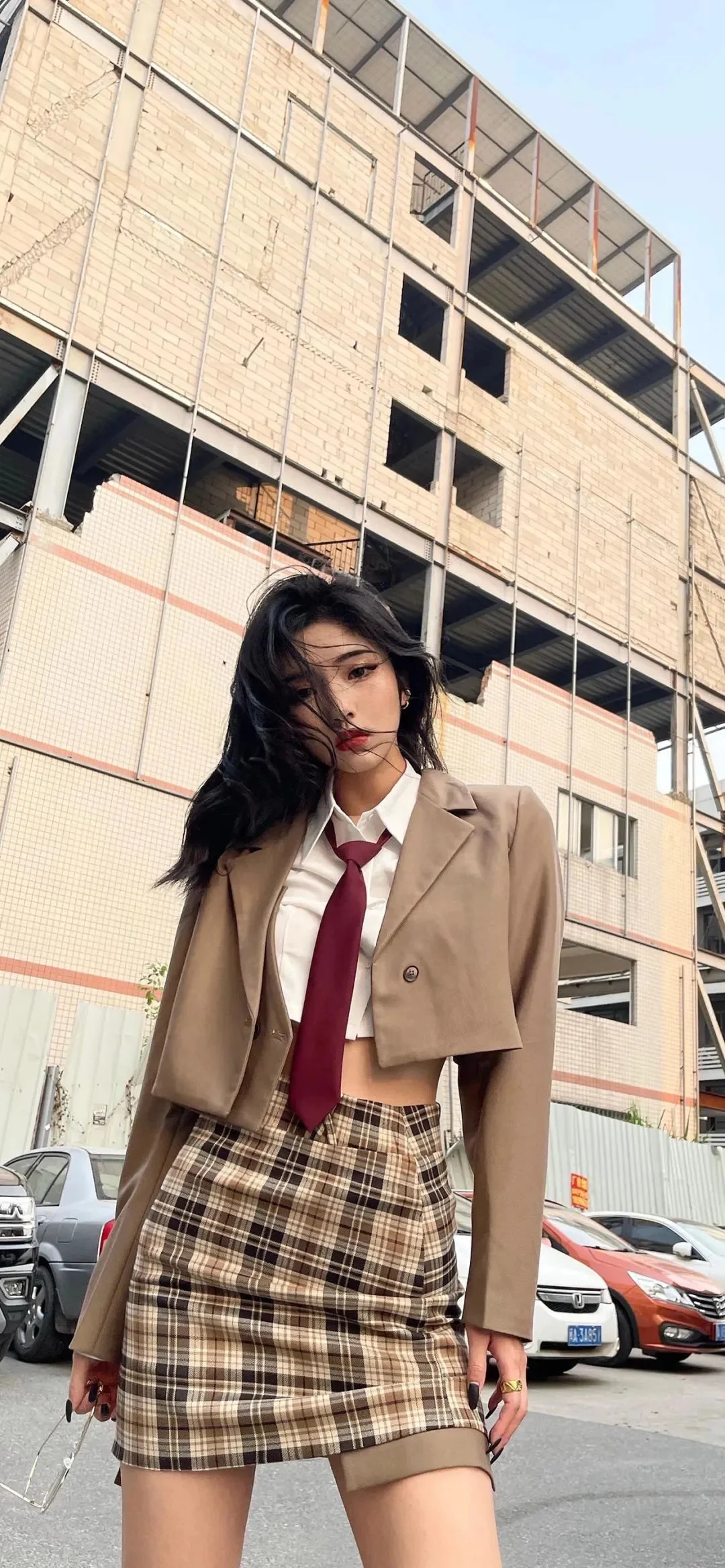 갈색 크롭 재킷과 흰 셔츠, 넥타이, 체크무늬 스커트를 입은 여성이 도심 건물 앞에 서 있다.