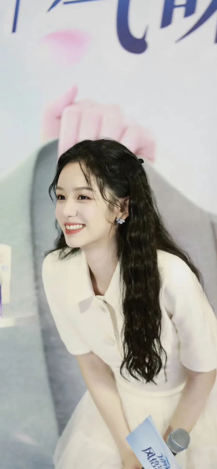 一位长发女性穿着白色上衣，面带微笑站在室内背景前。