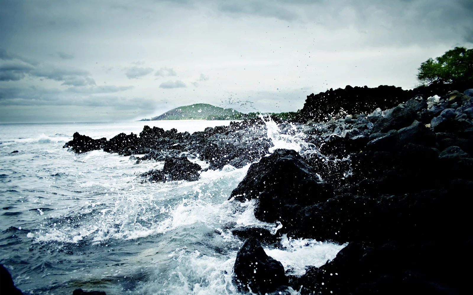 海浪衝擊黑色礁石海岸，遠處可見一座綠色小島。