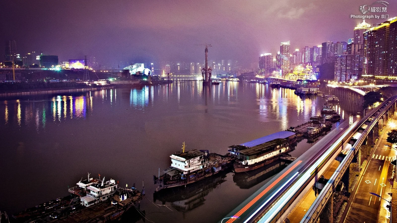夜色中的城市沿江而立，河面倒映着五彩灯光，前景道路上的车流形成明亮光轨。