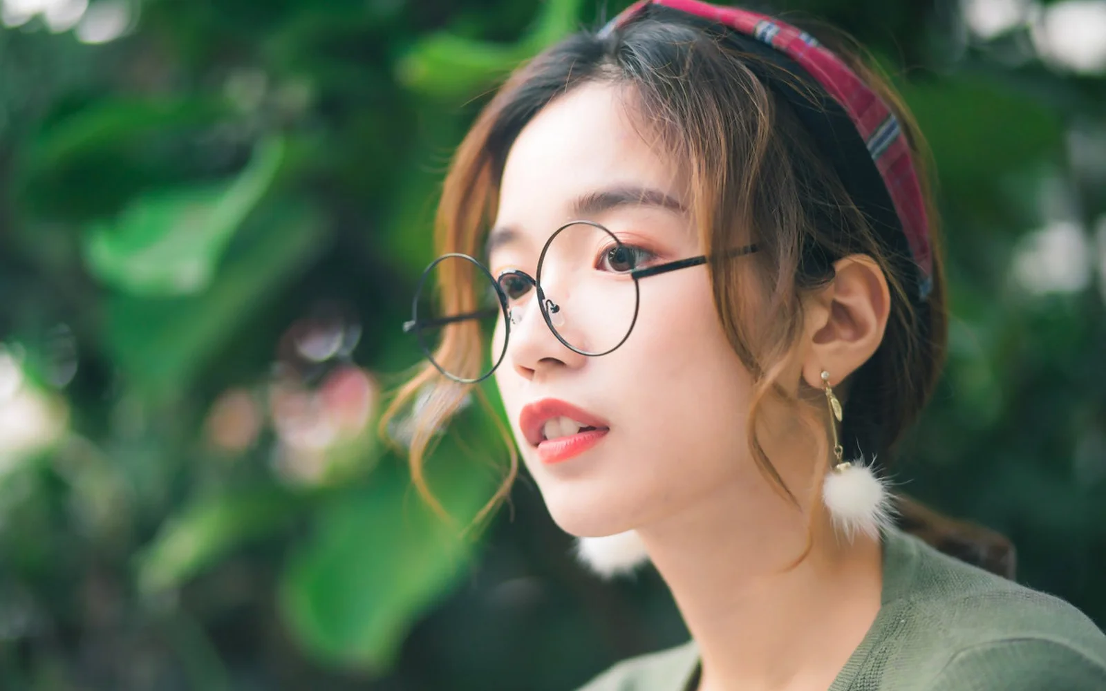 一位戴圆框眼镜的女性在绿叶背景前侧脸望向一旁。