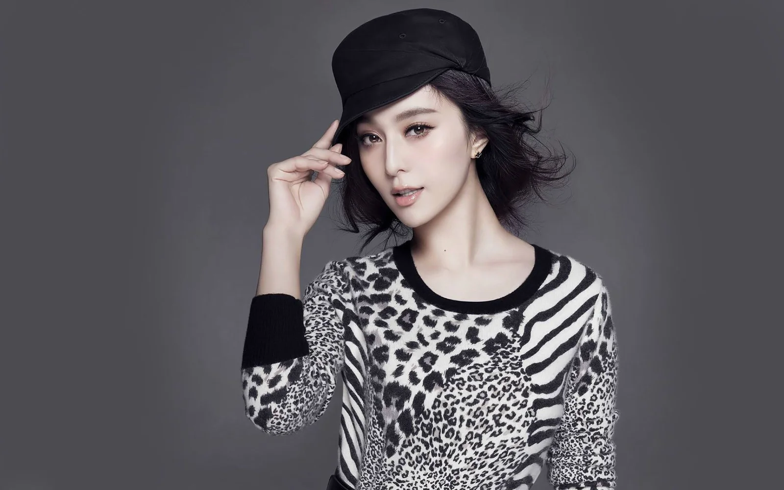 검은 모자와 애니멀 프린트 상의를 입은 성인 여성이 회색 배경 앞에서 포즈를 취하고 있다.