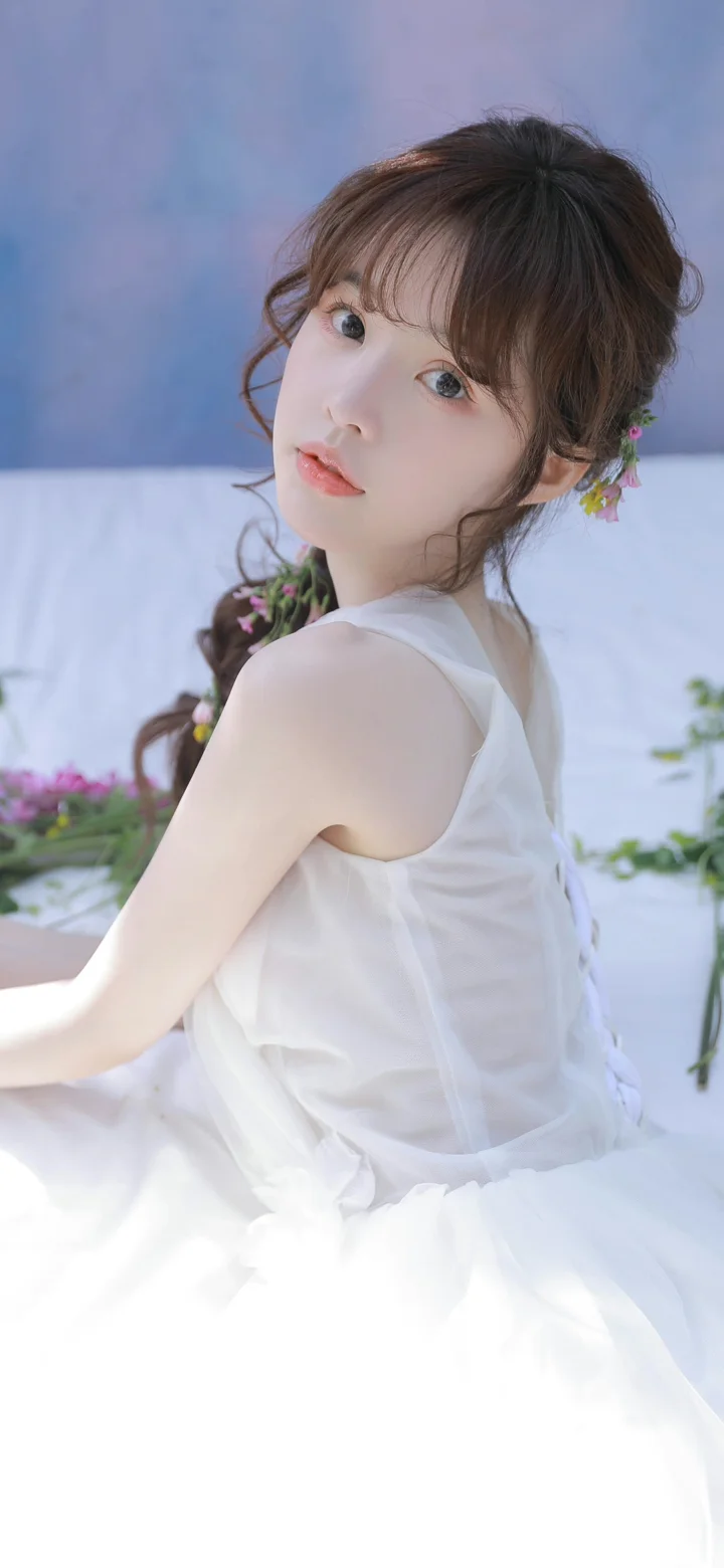흰색 민소매 드레스를 입은 여성이 옅은 파란색과 흰색 배경의 꽃 장식 앞에서 뒤돌아 카메라를 바라보고 있다.