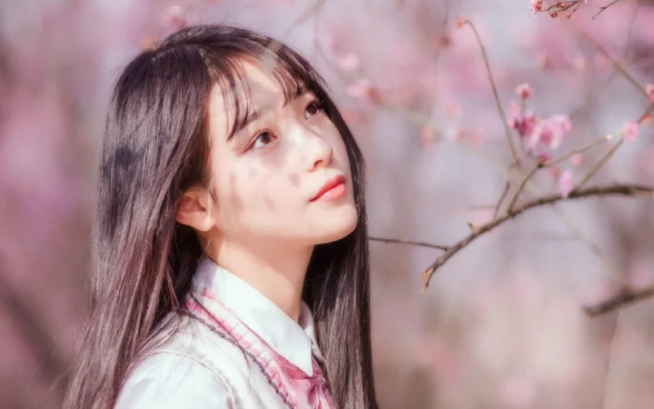 一名长发女子站在粉色樱花枝旁，柔和光线映衬着她的侧脸。