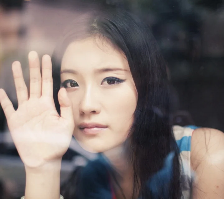 一名女性隔着玻璃抬手靠近脸部，呈现近距离肖像画面。