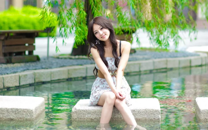 一名女子坐在池塘中的石台上，身后垂柳掩映，画面呈现出清新的户外氛围。