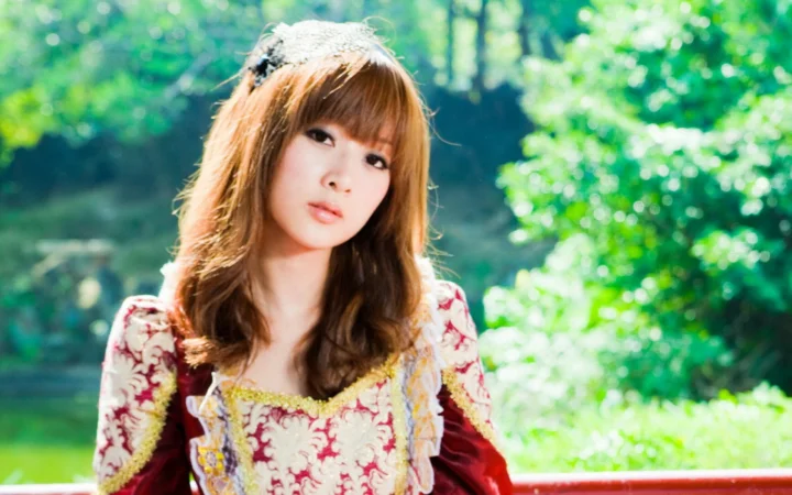 一名棕色长发女子站在阳光洒落的树林前，面向镜头微微侧头。