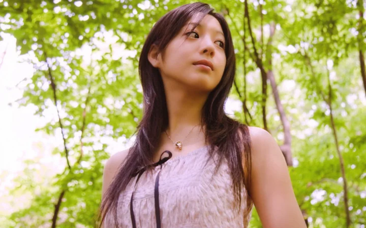 一名成年女性站在綠意盎然的森林中，陽光透過樹葉灑落在背景裡。