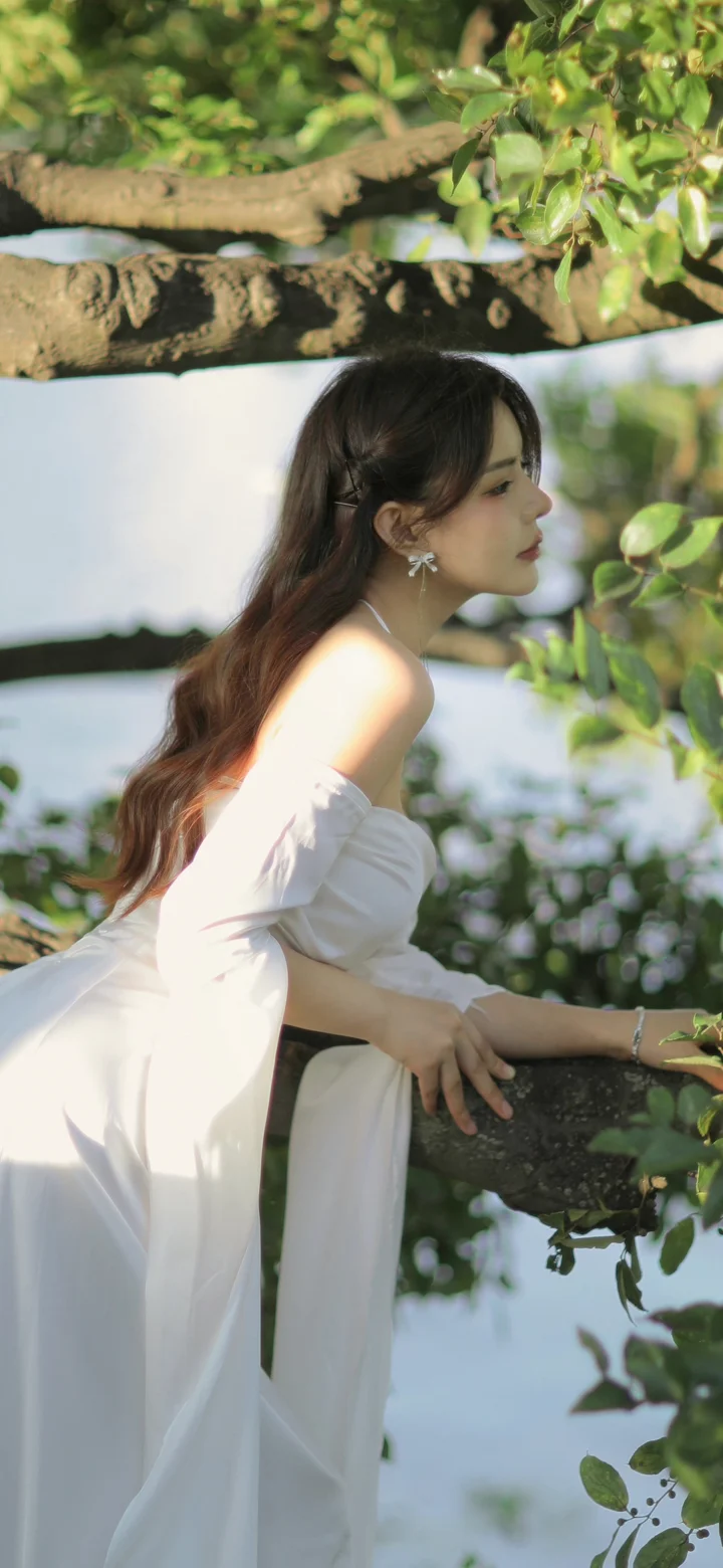 一名身穿白裙的女子倚靠在树干旁，周围是绿叶和柔和的自然光。