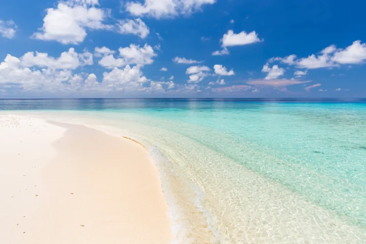 洁白沙滩延伸至清澈的 turquoise 海水，远处是平静海平面和蓝天白云。