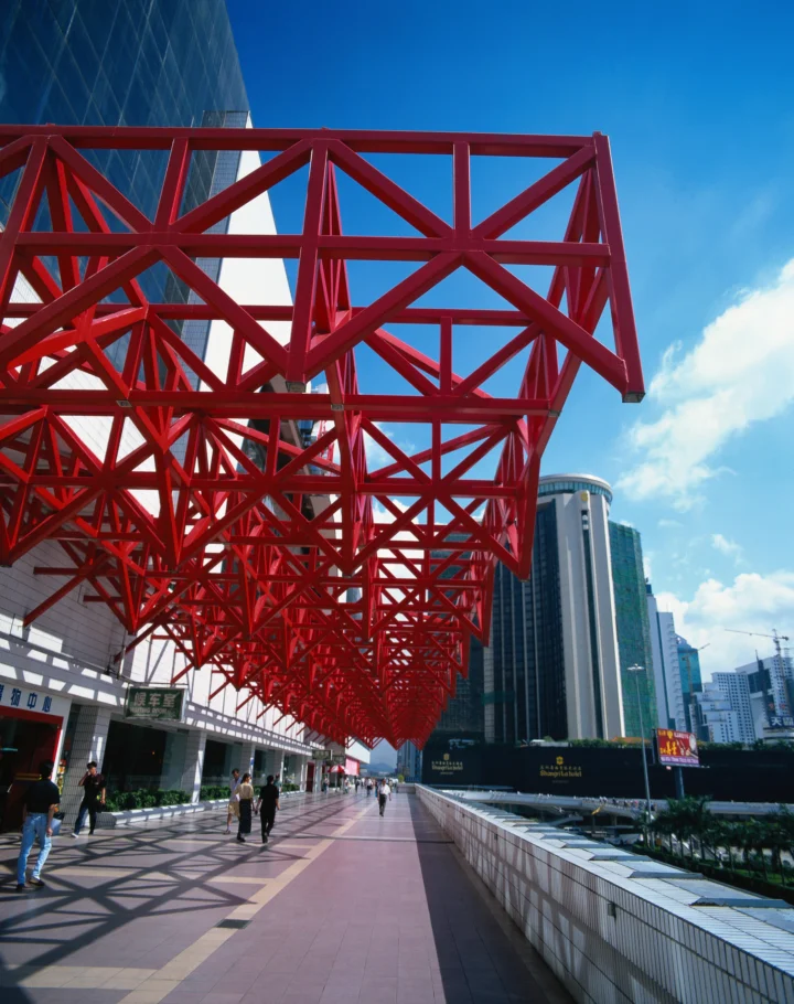 红色钢架结构横跨城市步道上方，周围可见现代高楼、蓝天和白云。