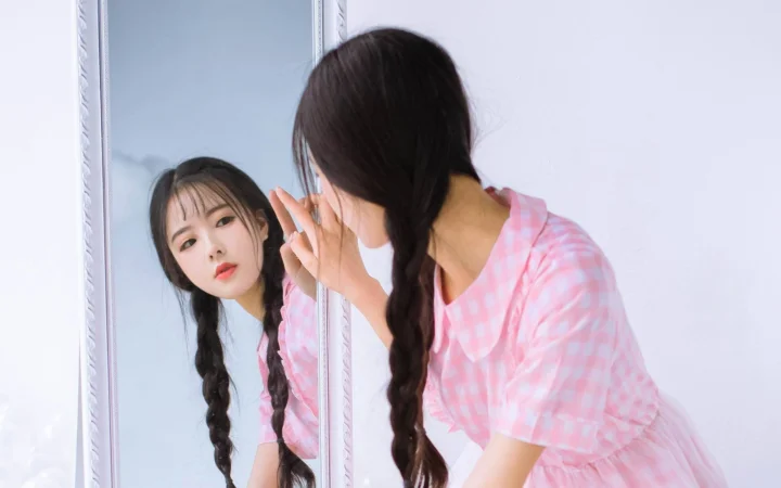 两名穿粉色衣服的女子站在镜子旁，其中一人通过镜面看向镜头。
