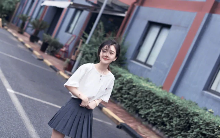 一名身穿白色上衣和深色裙子的女性站在城市街道上，身后是建筑和绿植。