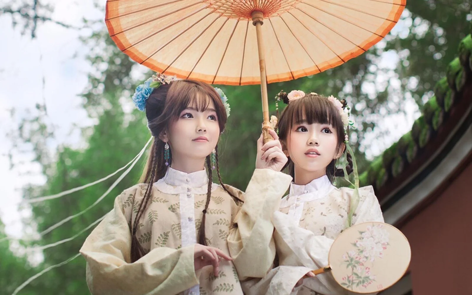 两位身穿浅色传统风格服装的女性站在户外树木间，一人撑着纸伞，另一人手持圆扇。