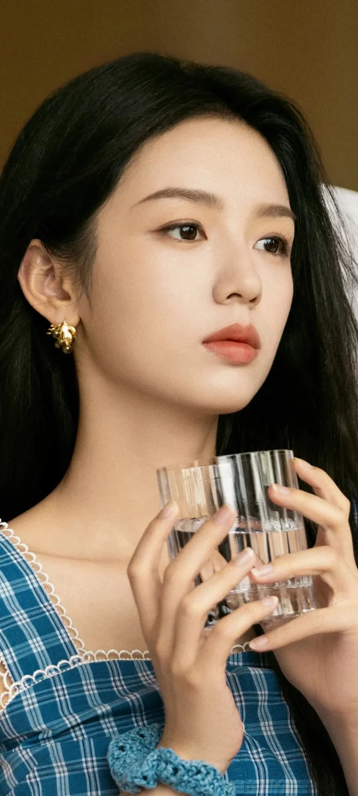 一位黑发女性身穿蓝色格纹上衣，手持透明玻璃杯面对镜头侧前方。