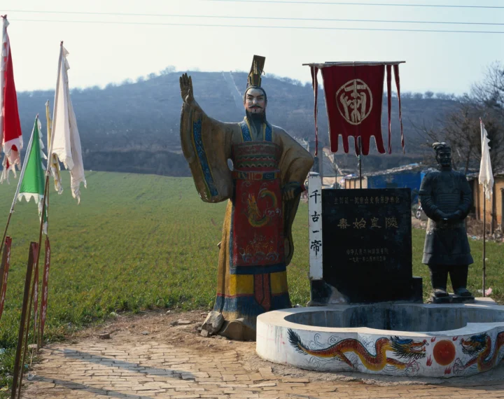 户外古迹中，一尊彩绘古代人物雕像、石井和彩旗映衬着远处的山坡。