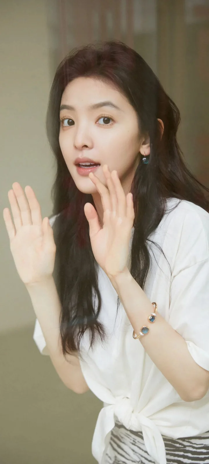 一名長髮女子穿著白色襯衫，在室內抬手靠近臉部面對鏡頭。