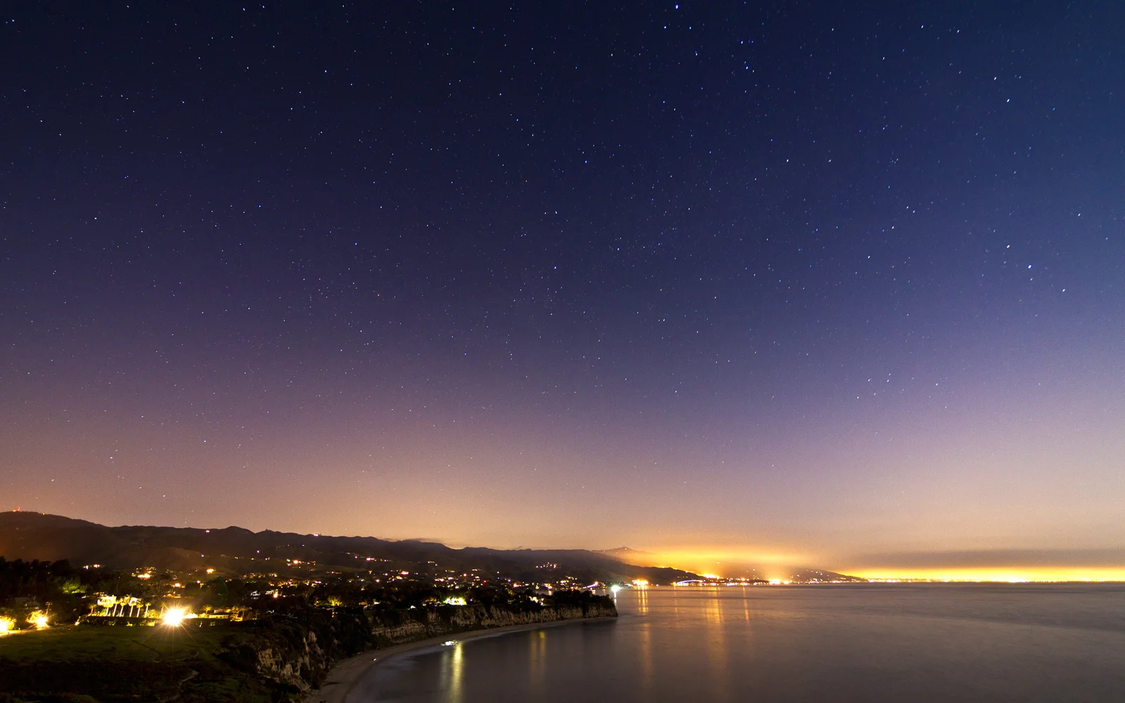 繁星点缀的夜空下，海湾、山丘和沿岸灯光倒映在平静的海面上。