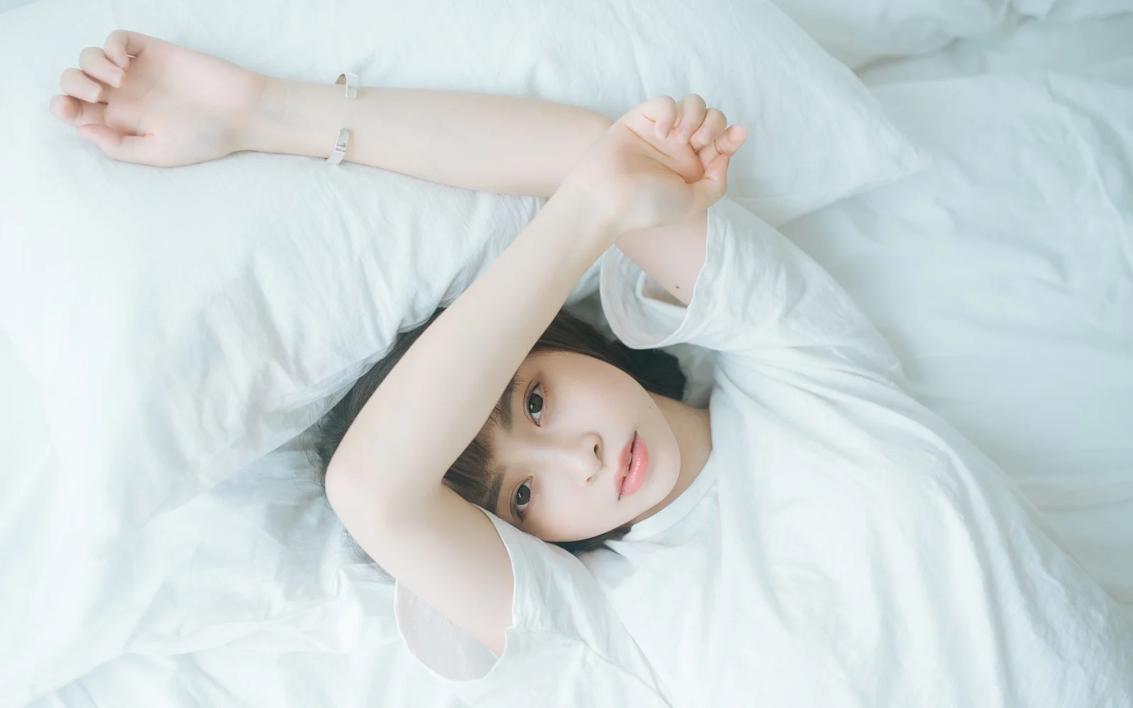 一名成年女性躺在白色床品上，双臂交叠在头顶。