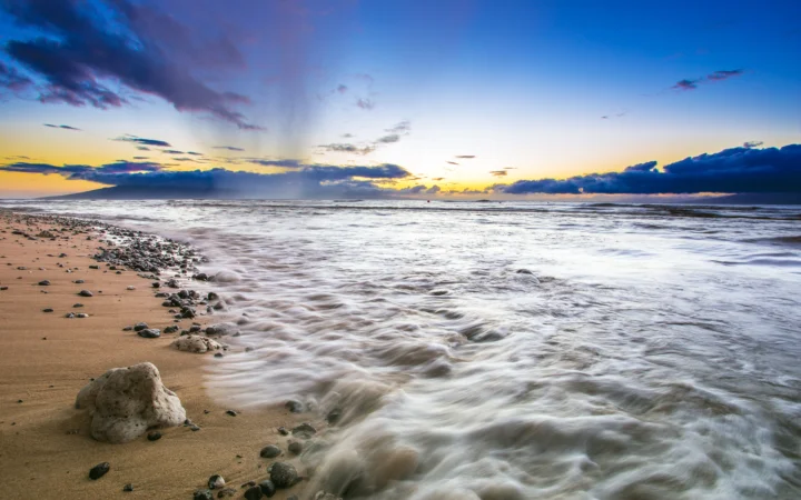 夕阳映照着海面，沙滩、礁石、翻涌的海浪与云层构成宁静的海岸风景。