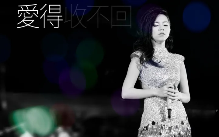 一名女性歌手身穿闪亮礼服，手持麦克风站在昏暗舞台上，背景有彩色虚化灯光。