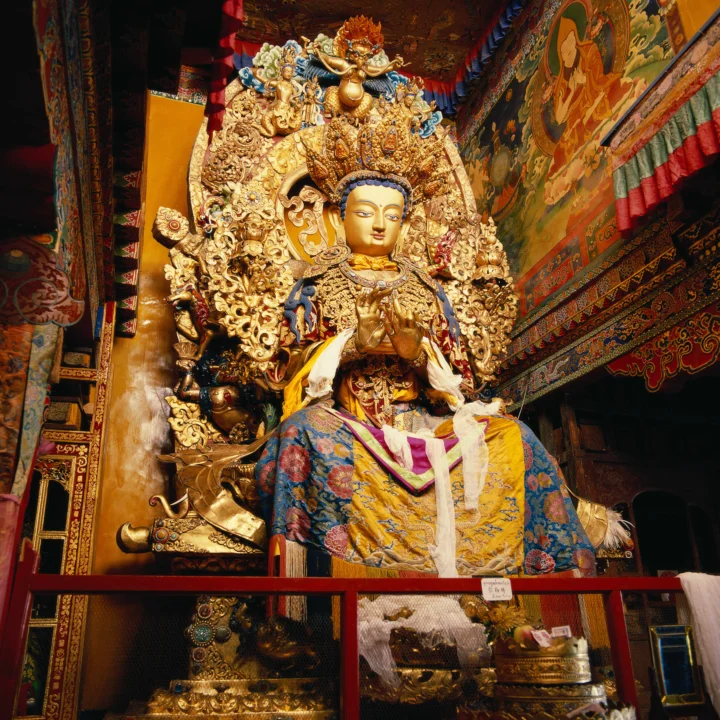 一尊身披彩色织物的金色佛像坐在装饰华丽的寺庙内部，周围环绕着精美雕刻和彩绘壁画。