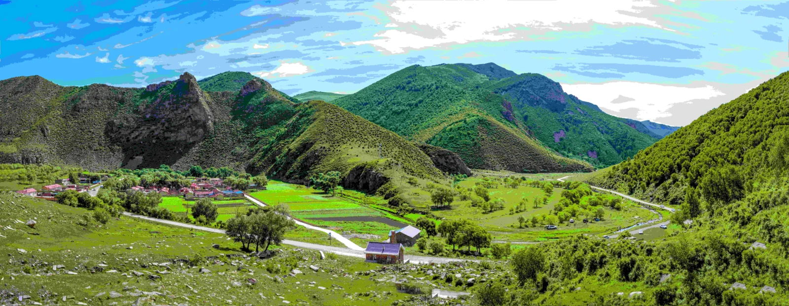 青翠群山环绕着山谷中的村庄、农田和蜿蜒道路，天空晴朗明亮。